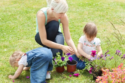 A woman with short blonde hair, wearing a light blue sleeveless top and dark jeans, is kneeling on a well-maintained lawn in a garden in Gipsy Hill. She is engaged in planting or tending to flowers, with her hands near a small potted plant. Beside her, there are additional pots with blooming plants, including purple and pink flowers. Two young children, a girl with light brown hair in a white T-shirt and blue jeans and a younger boy with curly hair in a blue dress, are playing on the grass and examining the flowering plants, with the boy bent over close to the ground. The garden features a lush green lawn, bordered by flower beds with vibrant, colorful flowers and plants, and has a backdrop of hedges and possibly trees. The scene appears to be sunny and bright, indicative of warm weather. The setting reflects typical outdoor gardening activity in a residential garden in Gipsy Hill, with a focus on lawn care and flower planting, highlighting the natural beauty of well-tended garden spaces.