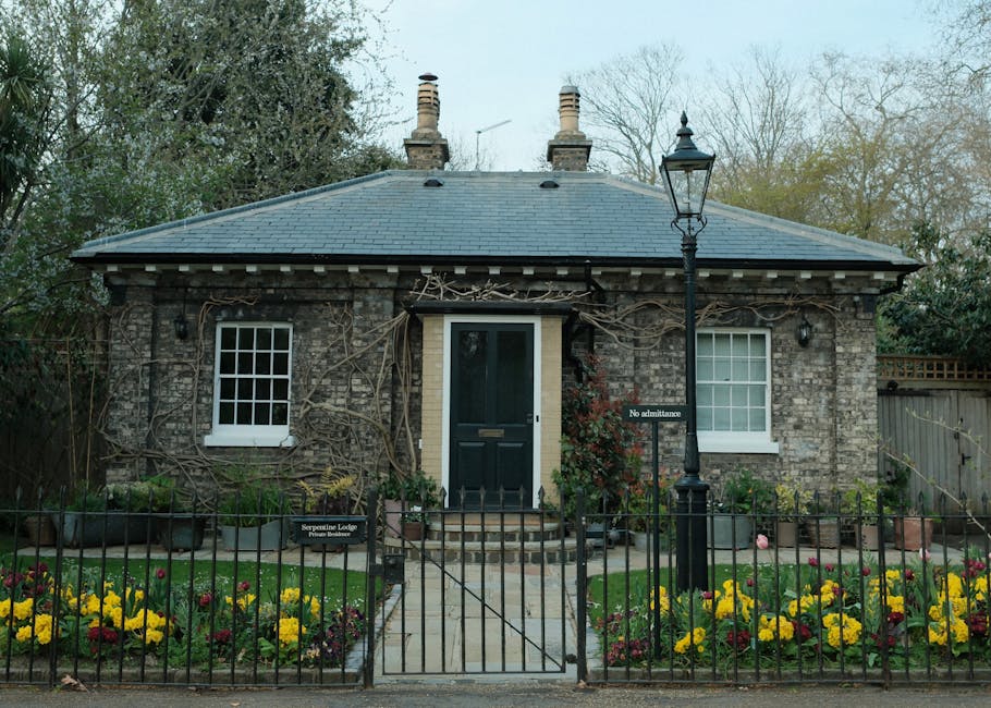 A small brick garden outbuilding situated behind a black iron fence, featuring two white-framed sash windows and a black central door with a letterbox, set within a landscaped garden. The garden has a