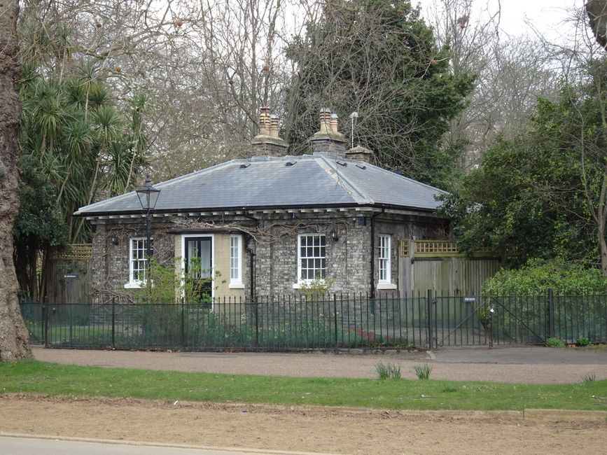 A small brick garden outbuilding situated behind a black iron fence, featuring two white-framed sash windows and a black central door with a letterbox, set within a landscaped garden. The garden has a